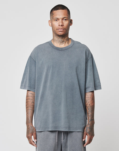 A man with short hair and visible tattoos on his neck and arms stands facing forward, wearing a loose-fitting LIVE FAST DIE YOUNG Basic Light Tee and matching gray pants against a plain light background.