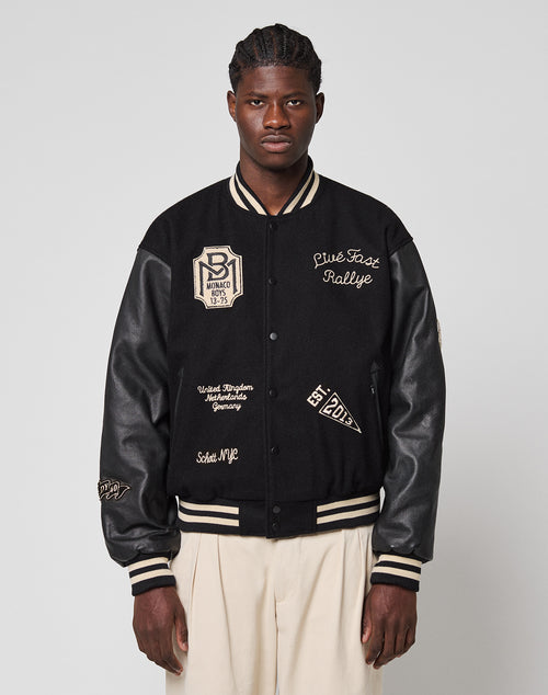 A man in cream pleated trousers wears the LFDY x Schott NYC Varsity Jacket by LIVE FAST DIE YOUNG, featuring black fabric with cream patches and writing, as he stands against a plain light gray background with a neutral expression.