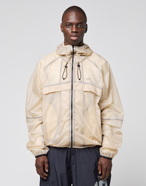 A man with short hair and a neutral expression wears the LIVE FAST DIE YOUNG Live Fast Nylon Jacket in light beige, featuring a front zipper, black drawstrings, and reflective print. He stands against a plain light gray background with his hands by his sides.