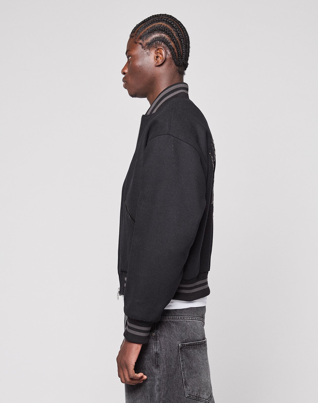 A person with braided hair, wearing the Live Fast College Jacket by LIVE FAST DIE YOUNG and dark gray jeans, stands in profile against a plain light gray background.