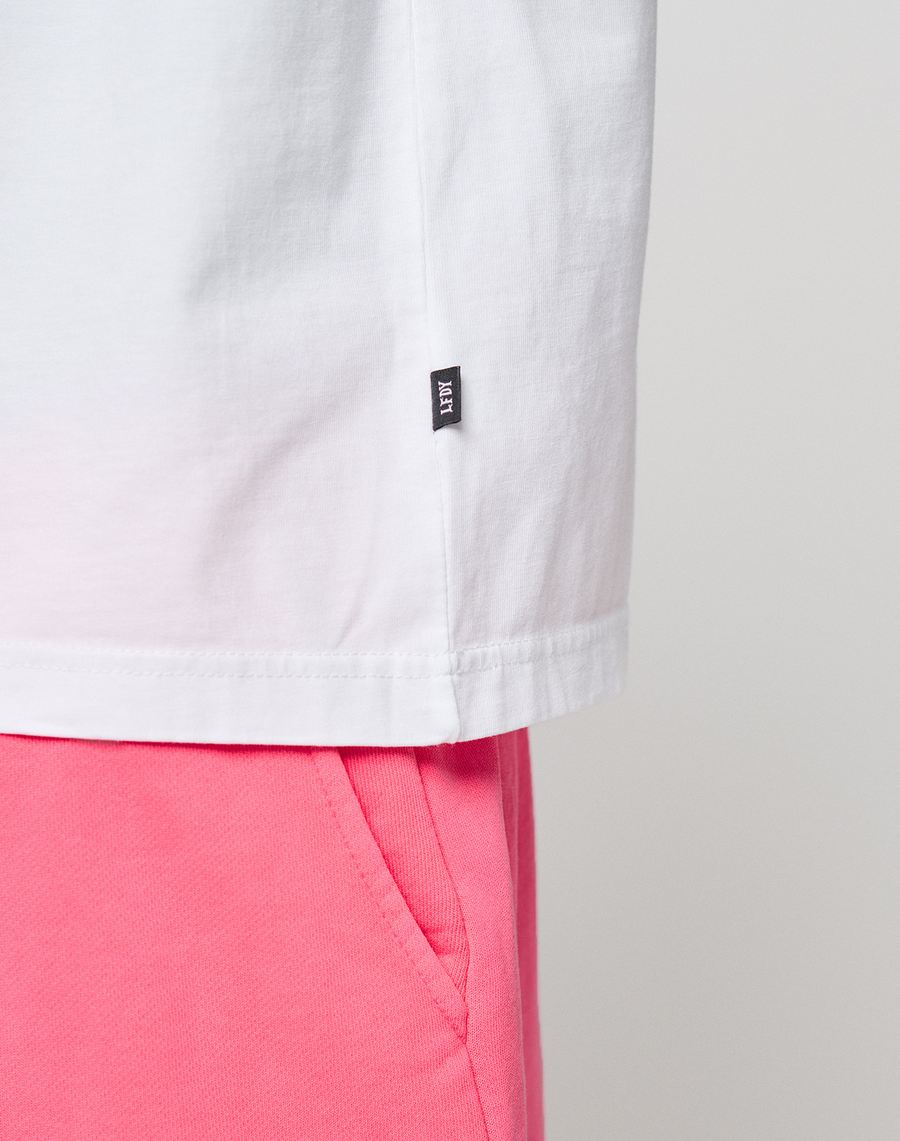 Close-up of the lower half of a white LIVE FAST DIE YOUNG Basic OS Livefast Tee (100% Baumwolle) with a small black tag, paired with bright pink shorts. The background is plain and light-colored.