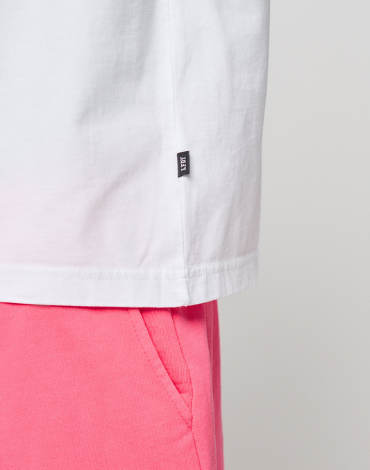 Close-up of the lower half of a white LIVE FAST DIE YOUNG Basic OS Livefast Tee (100% Baumwolle) with a small black tag, paired with bright pink shorts. The background is plain and light-colored.