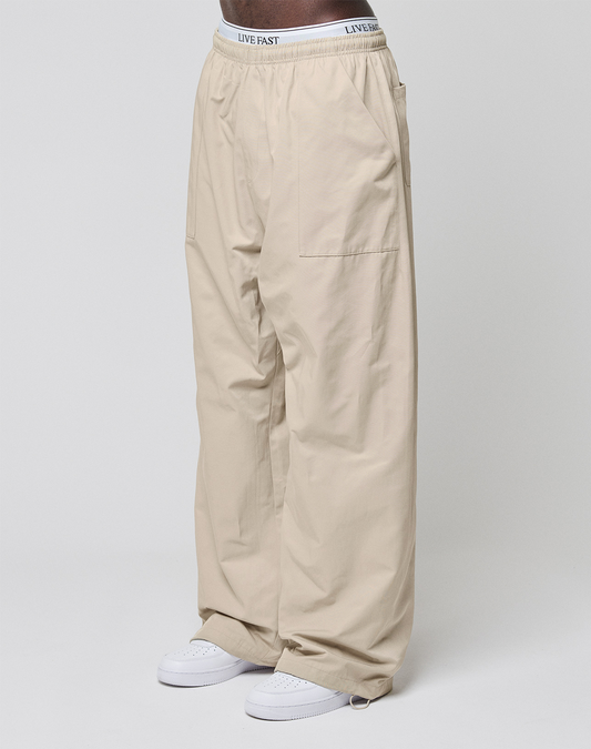 A person in Baggy Nylon Pants by LIVE FAST DIE YOUNG, featuring aufgesetzte Taschen and a white Elastikbund with "LIVE FAST," wears white sneakers against a plain light background.