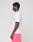 A man with short curly hair stands in profile, wearing a LIVE FAST DIE YOUNG Basic OS Livefast Tee in white with pink frontprint lettering and pink shorts, against a plain light gray background.