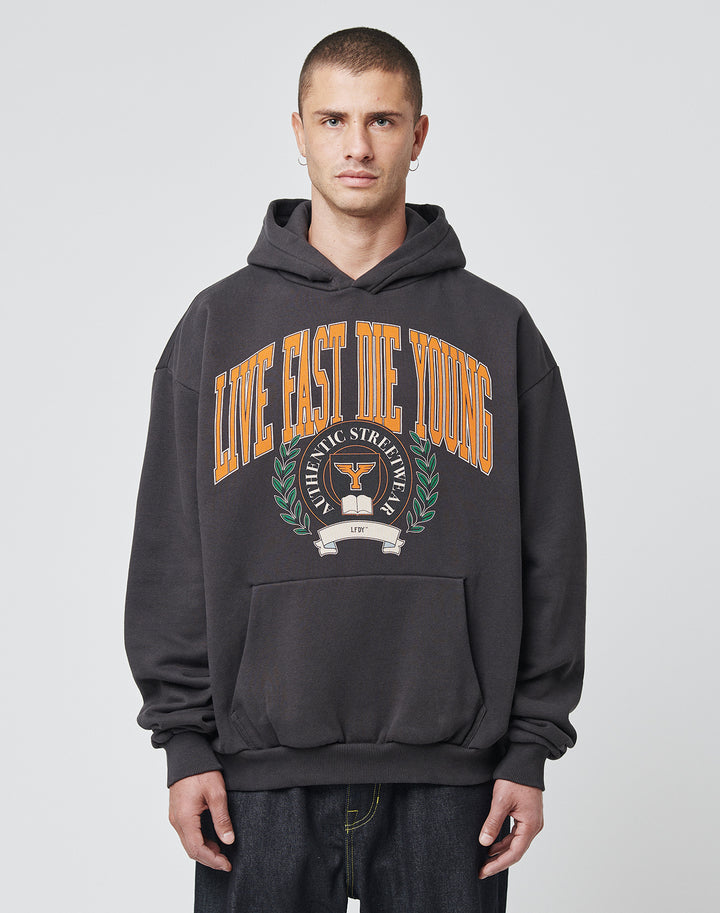 A young man with short hair wears the LIVE FAST DIE YOUNG Live Fast Die Young Hooded, a black hoodie with bold orange “LIVE FAST DIE YOUNG” lettering and laurel graphic, made from soft Baumwolle Polyester, standing before a plain light gray background. | model