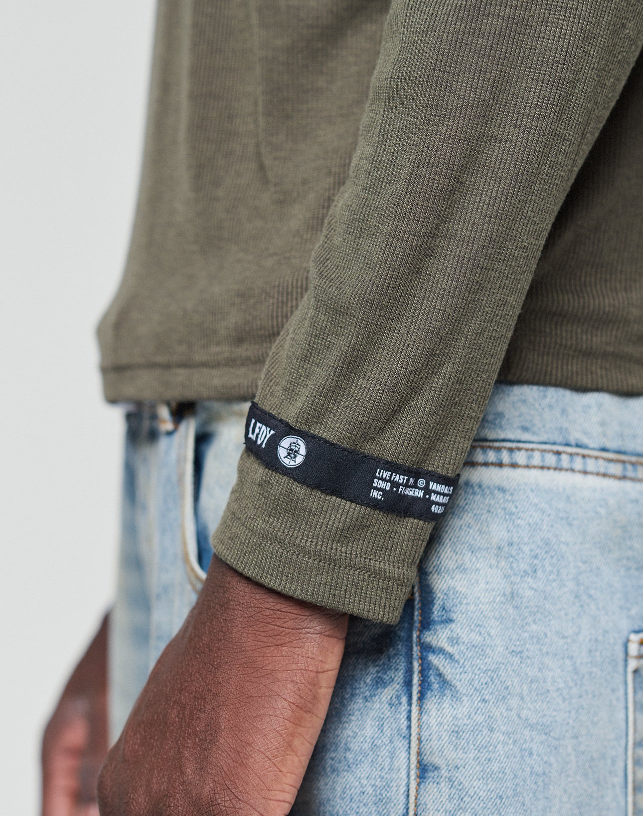 A close-up of an arm wearing the LIVE FAST DIE YOUNG LFDY Rib Longsleeve in olive green (220 GSM, ribbed, black and white web-label on cuff), paired with light blue denim jeans against a plain, light background.