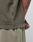 A close-up of a person in the LIVE FAST DIE YOUNG Basic +49 Chest Tee, an oversize greenish-brown shirt made from 100% Baumwolle, with a small black tag shown. Their tattooed arm and light mesh shorts are also visible.