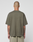 A person with a shaved head stands with their back to the camera, wearing an olive-green LIVE FAST DIE YOUNG Basic +49 Chest Tee and mesh shorts. Tattoos cover their forearms, set against a plain white background that highlights their style.