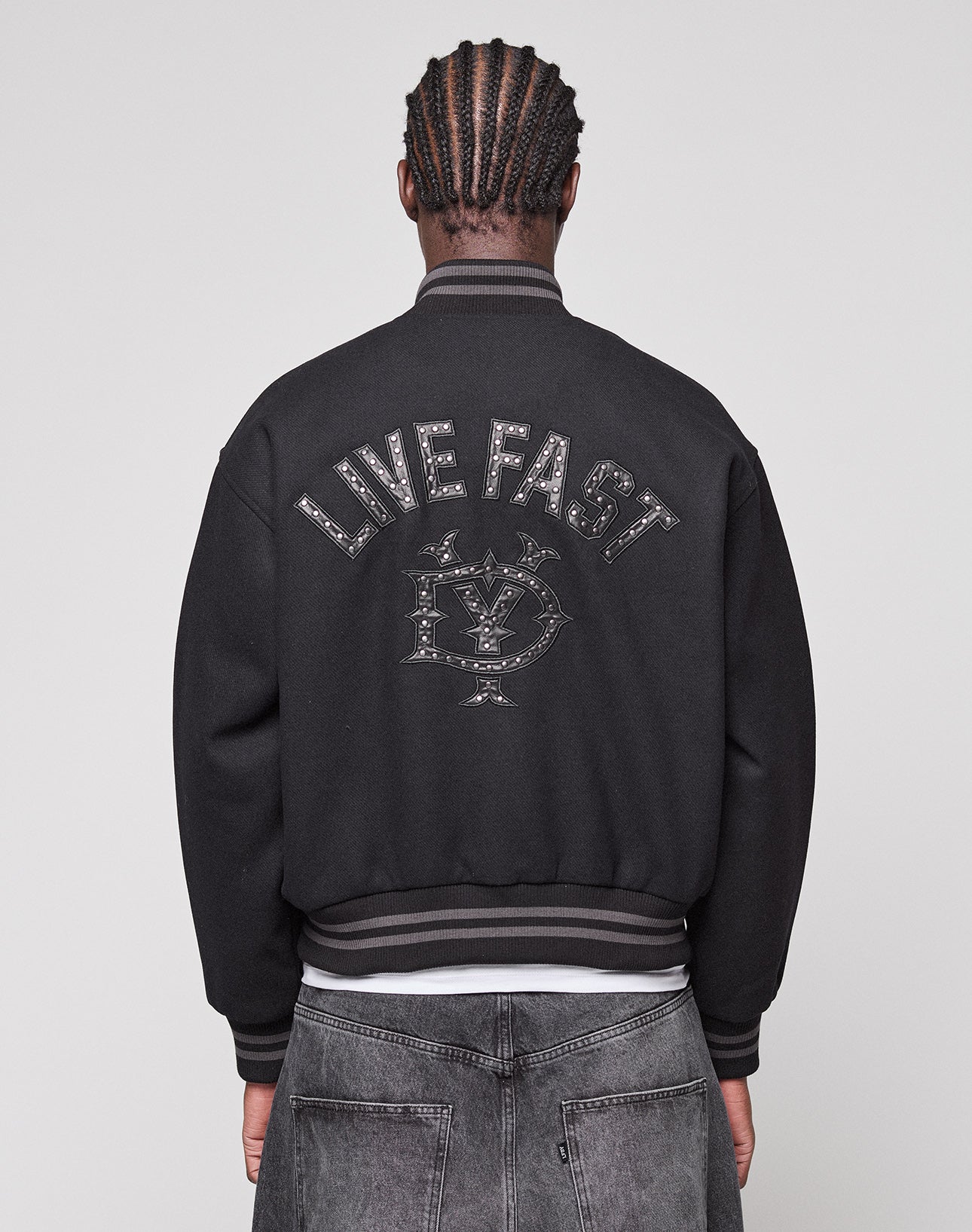 A person with braided hair is seen from behind, wearing the LIVE FAST DIE YOUNG Live Fast College Jacket with a Nieten Patch Logo on the back and metal zipper, paired with regular fit black jeans.