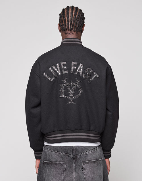 A person with braided hair is seen from behind, wearing the LIVE FAST DIE YOUNG Live Fast College Jacket with a Nieten Patch Logo on the back and metal zipper, paired with regular fit black jeans.