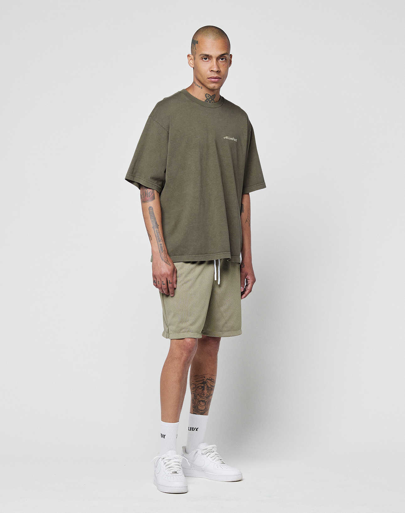 A tattooed person wears the LIVE FAST DIE YOUNG Basic +49 Chest Tee in olive green with matching shorts, white crew socks, and white sneakers, standing calmly against a plain white background.