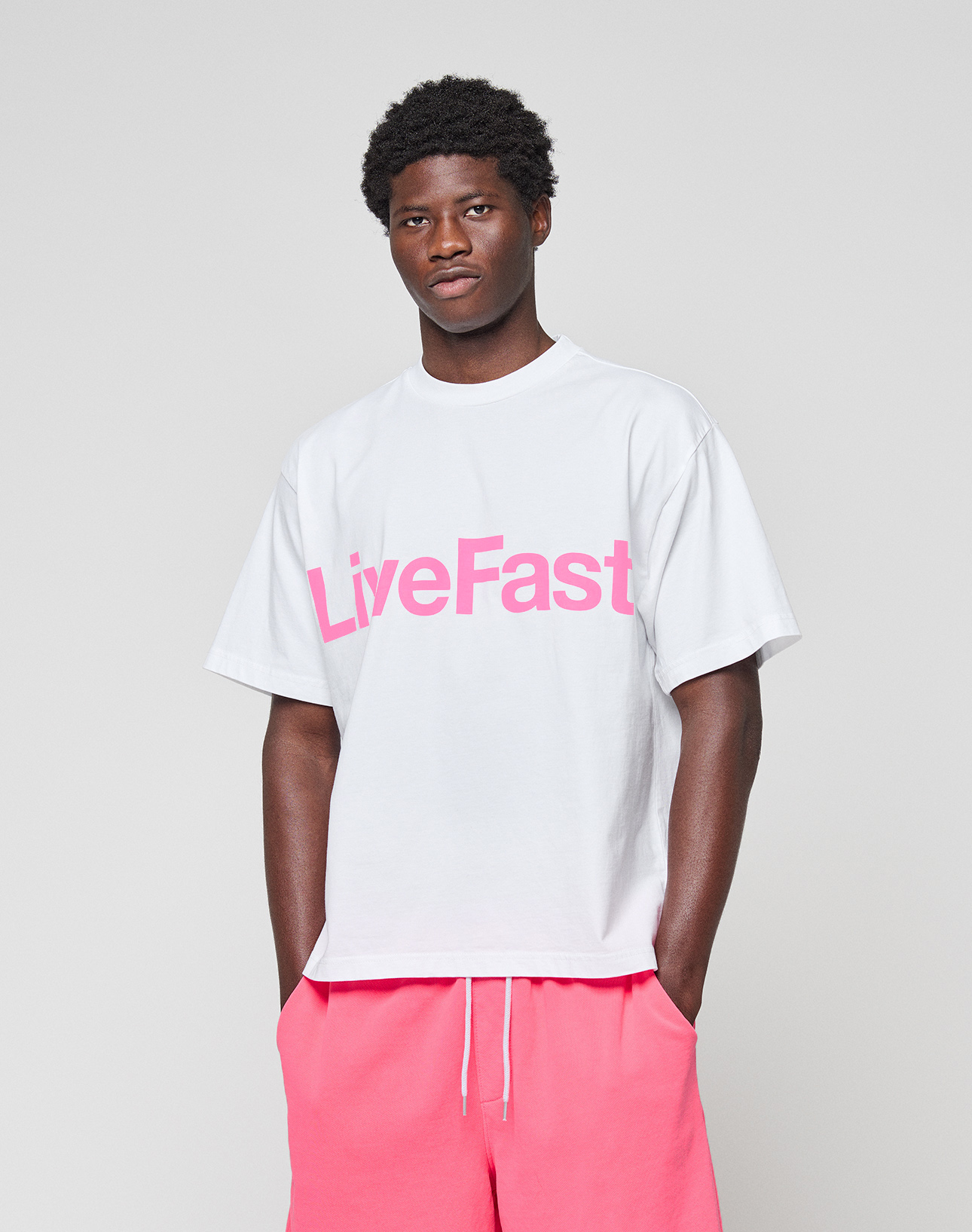 A man wears the Basic OS Livefast Tee by LIVE FAST DIE YOUNG, a white oversize fit t-shirt with a large pink "LiveFast" frontprint, paired with pink shorts, standing against a plain light gray background.
