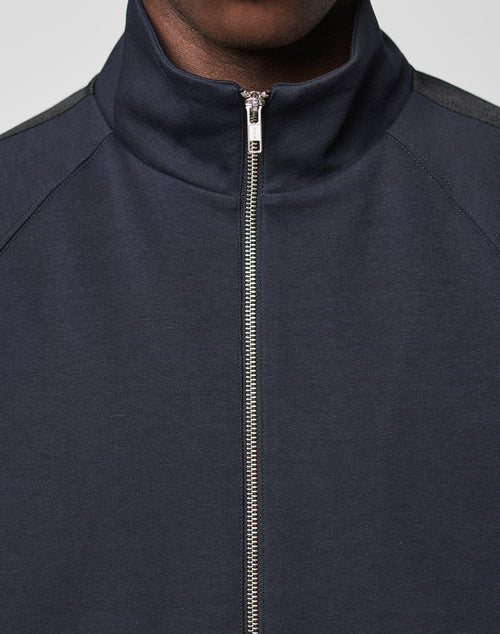 A close-up shows a person wearing the LFDY Striped Track Jacket by LIVE FAST DIE YOUNG, featuring a dark navy color, high collar, raglan sleeves, and a centered silver zipper. Only the lower half of the face and upper chest are visible.
