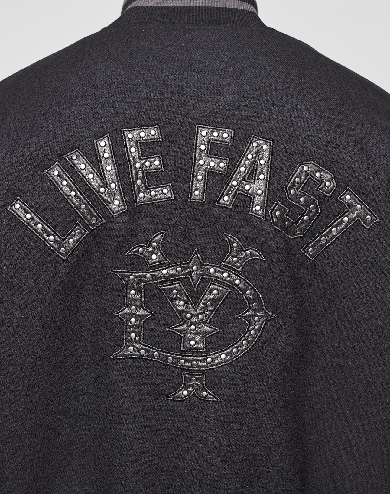 Close-up of the back of the Live Fast College Jacket by LIVE FAST DIE YOUNG, showing "LIVE FAST" in bold metal studs and a Nieten Patch Logo below, also accented with metal studs.
