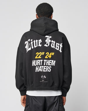 A person wears the LFDY Haters Hooded Zip by LIVE FAST DIE YOUNG, an oversize black hoodie with a double-layer hood and white/yellow Live Fast and 22 24 HURT THEM HATERS text plus small graphics on the back.