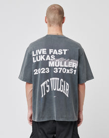 A person with short hair wears the LIVE FAST DIE YOUNG Its Vulgar Tee, a faded black oversized shirt featuring bold front and back prints. The back displays: LIVE FAST™ LUKAS MULLER 2023 370x51 ITS VULGAR in large white text. | model
