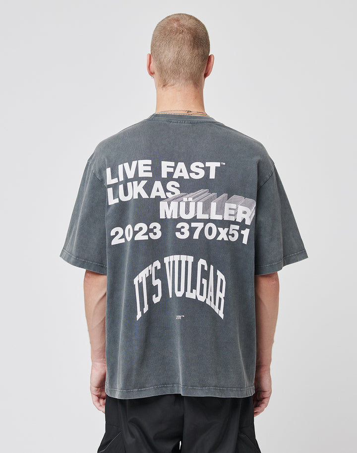 A person with short hair wears the LIVE FAST DIE YOUNG Its Vulgar Tee, a faded black oversized shirt featuring bold front and back prints. The back displays: LIVE FAST™ LUKAS MULLER 2023 370x51 ITS VULGAR in large white text. | model