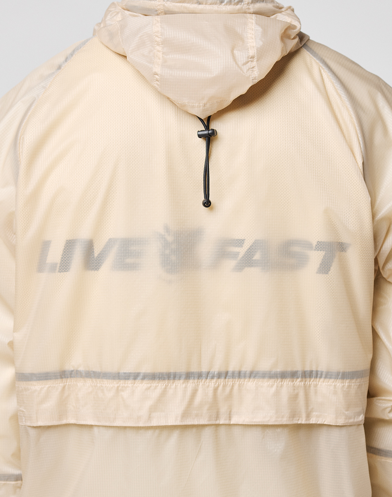 A close-up shows the back of the Live Fast Nylon Jacket by LIVE FAST DIE YOUNG, featuring a reflective "LIVE FAST" print in bold letters and a black drawstring beneath the hood, accentuating its light beige transparent nylon fabric.