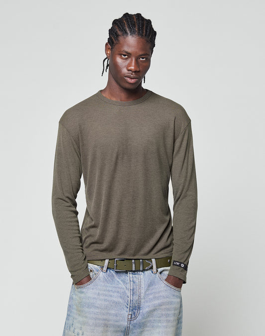 A man with braided hair stands with his hands in his pockets against a plain light gray background, wearing an olive green LIVE FAST DIE YOUNG LFDY Rib Longsleeve, light blue jeans, and a black belt.
