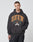 A man wears the LIVE FAST DIE YOUNG Live Fast Die Young Hooded with text and crest graphic, made from Baumwolle Polyester. He stands against a plain light background, facing the camera with a neutral look and subtle vintage effect.