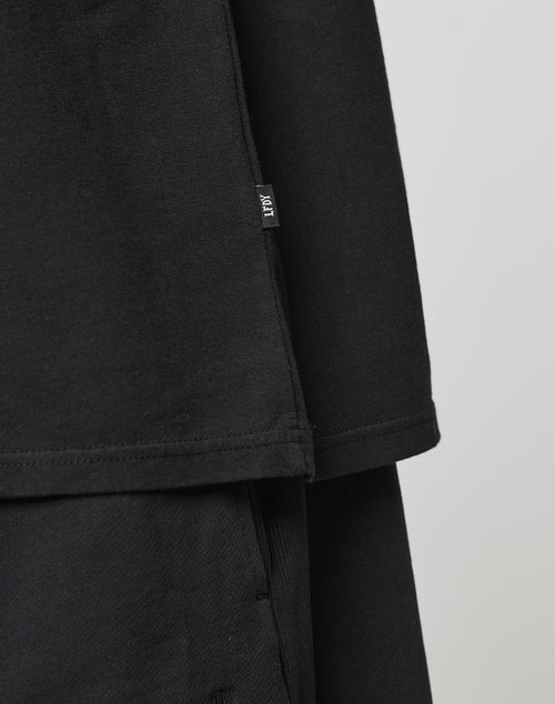 Close-up of the bottom half of a black Basic Muscle Shirt by LIVE FAST DIE YOUNG, styled over black pants. A small white-text fabric tag is sewn into the side seam. The background is plain and light-colored.