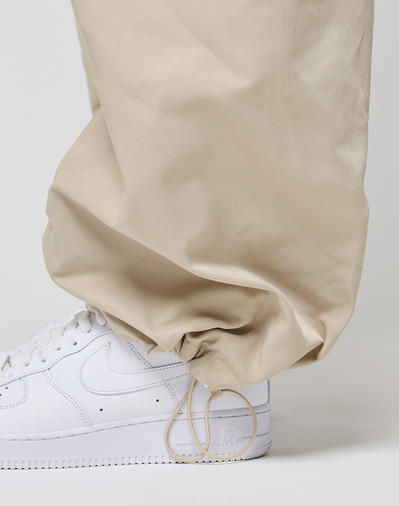 A close-up of a person wearing LIVE FAST DIE YOUNG Baggy Nylon Pants with a wide fit and unique ankle drawstrings, paired with white Nike Air Force 1 sneakers.