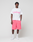 A man stands against a plain background wearing the LIVE FAST DIE YOUNG Basic OS Livefast Tee—an oversize fit, 100% Baumwolle T-shirt with bold pink frontprint—paired with bright pink shorts, white crew socks, and white sneakers.