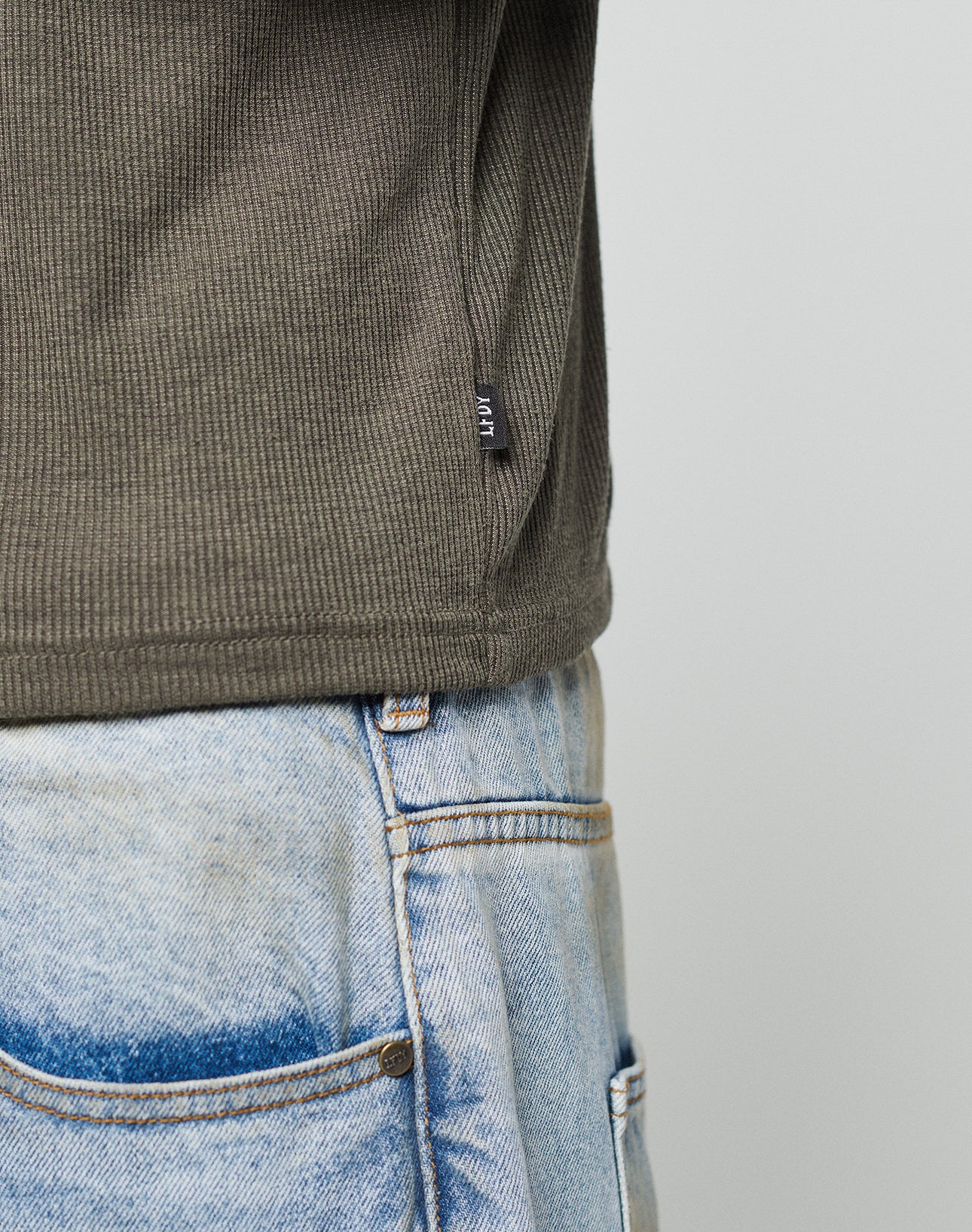 A close-up of a person wearing the LIVE FAST DIE YOUNG LFDY Rib Longsleeve in dark green and light blue denim jeans, highlighting the shirt’s subtle web-label on the side and the waistband and pocket of the jeans.