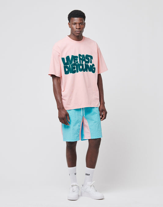 A man stands against a plain background wearing a pink LIVE FAST DIE YOUNG T-shirt, aqua blue and pink Beach Shorts from LIVE FAST DIE YOUNG with mesh inlay, white socks, and white sneakers.