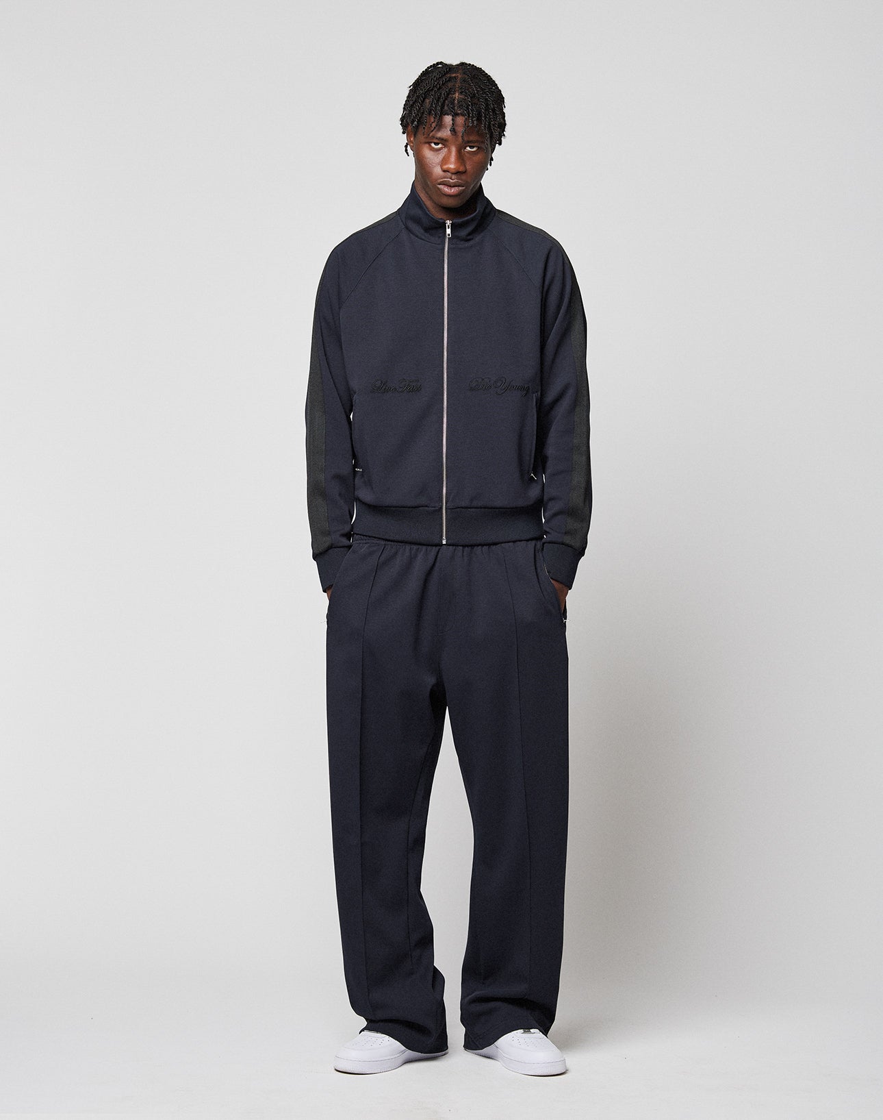 A man with short, twisted hair and a neutral expression faces forward against a plain light background, wearing the LIVE FAST DIE YOUNG LFDY Striped Track Jacket, matching wide-leg trousers, and white sneakers.