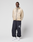 A person stands against a white background wearing a beige nylon hooded jacket, LIVE FAST DIE YOUNG LFDY Performance Trackpants in dark navy with an oversize fit and "40" on the left leg, and white sneakers. Their hands are behind their back.
