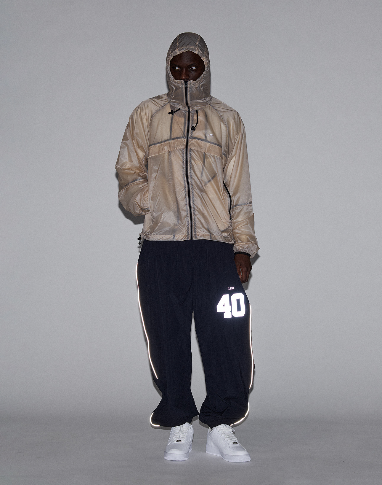 A person in a tan LIVE FAST DIE YOUNG Live Fast Nylon Jacket with transparent shell zipped up to their mouth, black pants featuring a reflective "40" print on one leg, and white sneakers stands against a plain gray background.