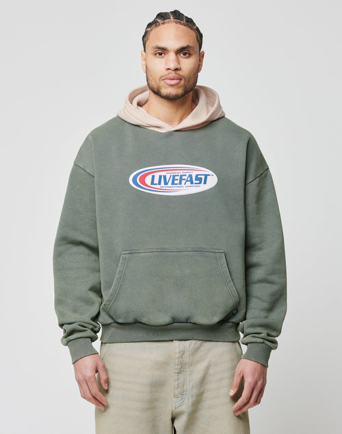 A man with braided hair wears the LIVE FAST DIE YOUNG Live Fast Garment Hooded, an oversized green hoodie with a beige hood and chest logo, standing against a plain light background.