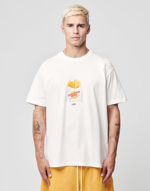 A person with short, bleached blonde hair wears a loose white LFDY Fries Tee by LIVE FAST DIE YOUNG, featuring a graphic of fries in a red carton labeled “Everybody Fries,” and yellow shorts, against a plain light gray background. LF2406-15695.
