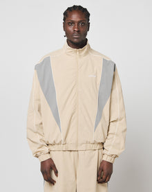 A man with braided hair wears the LIVE FAST DIE YOUNG Basic Nylon Trackjacket, a beige regular fit made of polyester and nylon with gray and white accents, standing against a light background facing the camera neutrally.