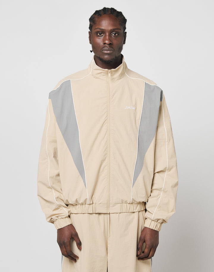 A man with braided hair wears the LIVE FAST DIE YOUNG Basic Nylon Trackjacket, a beige regular fit made of polyester and nylon with gray and white accents, standing against a light background facing the camera neutrally.