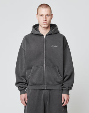 A person with short hair wears a dark gray LIVE FAST DIE YOUNG Basic Script Hooded Zip hoodie and matching sweatpants, standing against a plain light background and looking directly at the camera with a neutral expression.