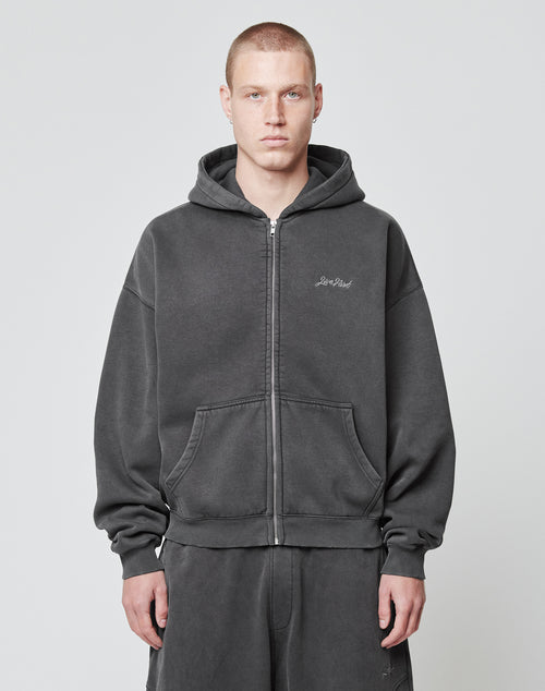 A person with short hair wears a dark gray LIVE FAST DIE YOUNG Basic Script Hooded Zip hoodie and matching sweatpants, standing against a plain light background and looking directly at the camera with a neutral expression.