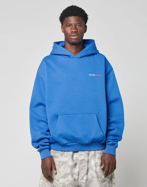 A man wearing an oversize fit LFDY F*ck Phones Hooded blue hoodie by LIVE FAST DIE YOUNG, made of soft polyester with a front pocket, and light gray patterned pants stands facing forward against a plain light background.