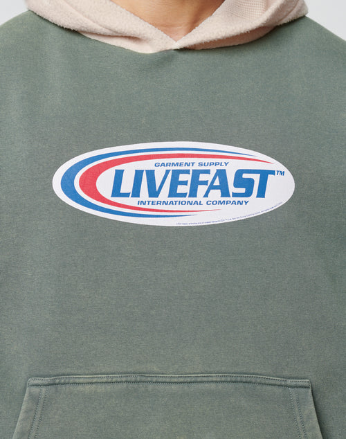 A close-up of the Live Fast Garment Hooded by LIVE FAST DIE YOUNG, an oversized green hoodie with a red and blue oval logo reading GARMENT SUPPLY LIVEFAST INTERNATIONAL COMPANY, front pocket, and cream hood lining.