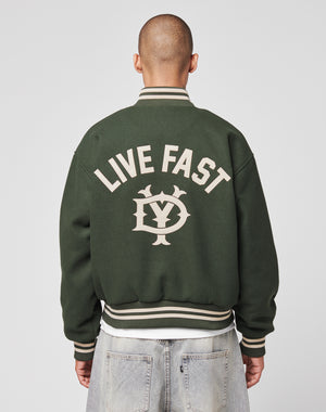 A person with a shaved head is seen from behind wearing the Live Fast College Jacket by LIVE FAST DIE YOUNG. The dark green varsity features white trim and large LIVE FAST text above a LY logo embroidered on the back.