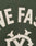 Close-up of green fabric with white stitched applique letters spelling NE FAS, ornate white design below, and a DY-Reißverschluss for contrast—details from the LIVE FAST DIE YOUNG Live Fast College Jacket.