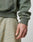 A close-up of a hand and lower arm wearing the LIVE FAST DIE YOUNG Live Fast Garment Hooded in green, paired with beige subtly striped pants, highlights the texture and relaxed fit of both pieces.