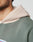 Close-up of a person wearing the soft, light beige Live Fast Garment Hooded by LIVE FAST DIE YOUNG under an oversized sage green sweatshirt. Only part of their face, neck, and shoulder are visible; the hoodie has a slightly fuzzy texture.
