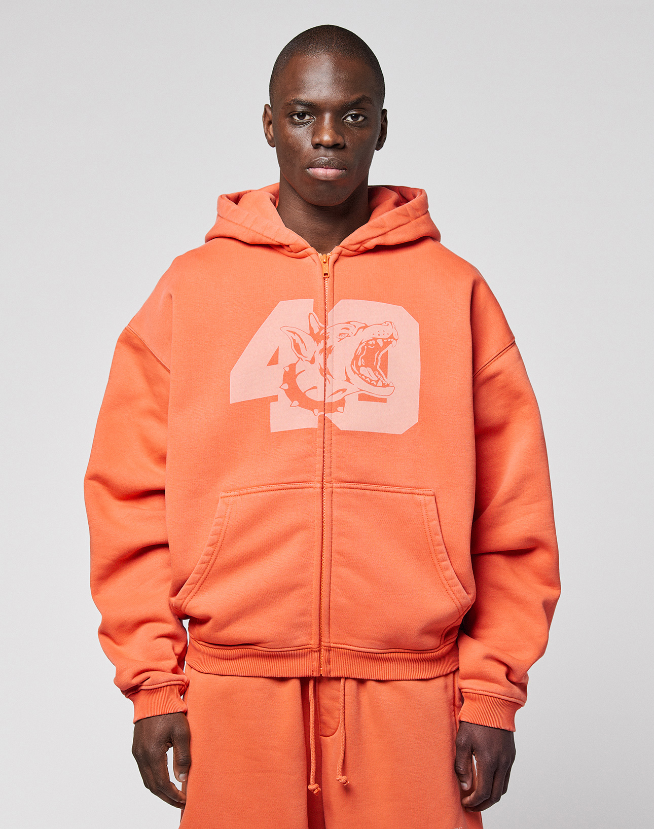 A person wearing the orange LFDY 40 Hooded Zip by LIVE FAST DIE YOUNG, featuring a large number 49 and shark front print, stands against a plain light gray background.