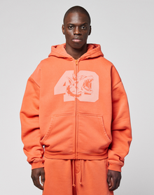 A person wearing the orange LFDY 40 Hooded Zip by LIVE FAST DIE YOUNG, featuring a large number 49 and shark front print, stands against a plain light gray background. | model