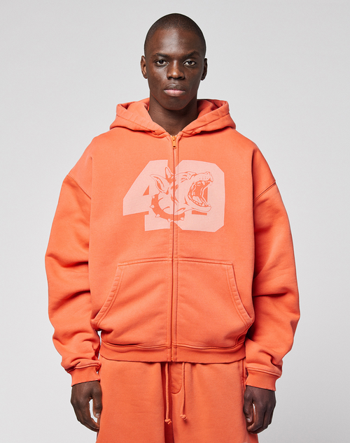 A person wearing the orange LFDY 40 Hooded Zip by LIVE FAST DIE YOUNG, featuring a large number 49 and shark front print, stands against a plain light gray background.