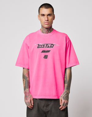 A man with tattoos wears the LFDY Triple Racing Tee by LIVE FAST DIE YOUNG—an oversize, bright pink 100% cotton T-shirt with a bold multilogo LIVE FAST front print—standing against a plain light gray background.