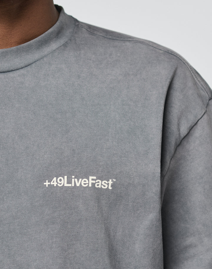 A person wears the LIVE FAST DIE YOUNG Basic +49 Chest Tee—an oversize gray shirt made from 100% Baumwolle, featuring a white +49LiveFast logo on the left chest. Only part of the upper body and shirt text are visible.