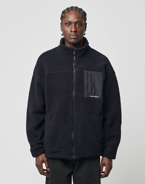 A man with braided hair models the LIVE FAST DIE YOUNG Basic Teddy Jacket, a black fleece zip-up with a stand-up collar and zippered chest pocket, against a plain light gray background, facing the camera.